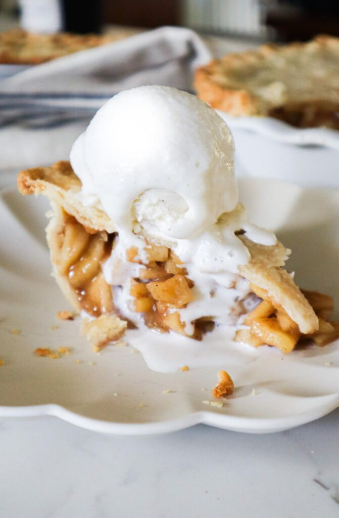 A slice of classic apple pie topped with a scoop of melting vanilla ice cream rests on a white plate. The pies flaky crust and generous apple filling are irresistible, with crumbs scattered around. Another tempting pie is blurred in the background.