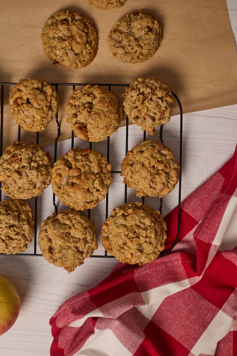 Oatmeal apple cinnamon cookies cooling on a rack with apples and a red checked towel.