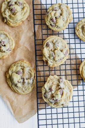 Freshly baked chocolate chip cookies are cooling on a black wire rack, with a few chilling on brown parchment paper on a white counter.