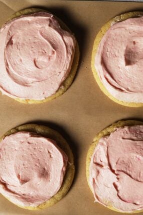 Four sugar cookies with pink frosting swirls sit on brown parchment paper, seen from above.