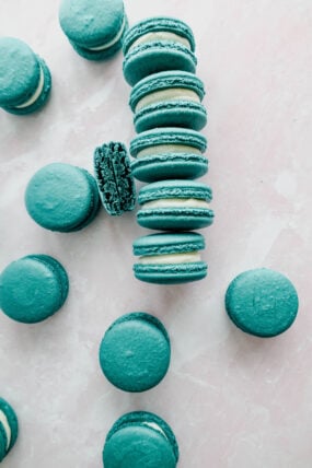 A bunch of blue-green macarons with creamy filling are scattered on a light pink marbled surface. Ones been bitten in half and is chilling there with the rest.