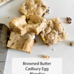 A stack of browned butter Cadbury egg blondies sits on a white surface, spatula and crumbs making it look extra tasty.