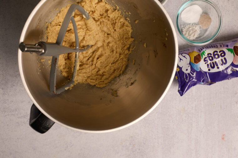 Mixer bowl with cookie dough and paddle, a small bowl of salt and baking powder, plus mini chocolate eggs on the counter—yum!.