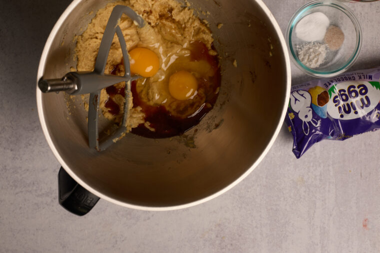 Top-down shot of a bowl of cookie dough, eggs, vanilla, mixer paddle, little bowls of dry stuff, and a bag of mini chocolate eggs.