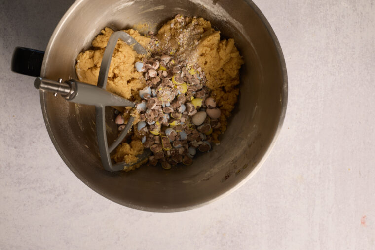 A metal bowl filled with cookie dough and crushed chocolate candies for blondies, with a paddle attachment ready to mix.