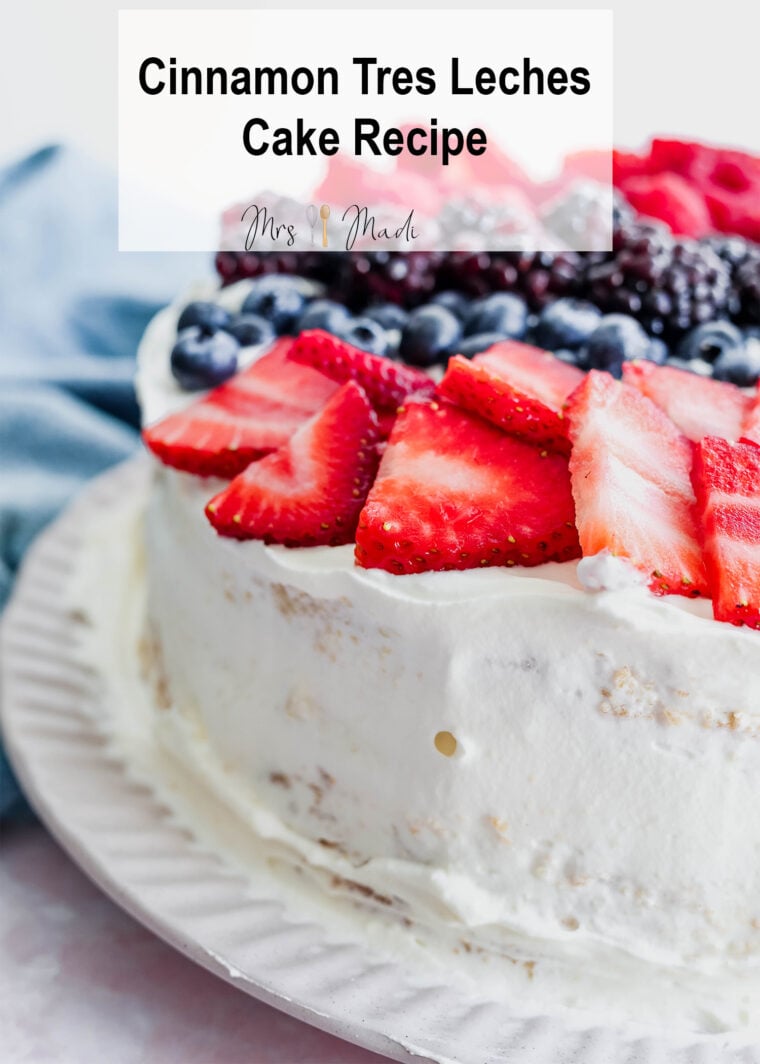A white-frosted tres leches cake loaded with fresh strawberries, blueberries, and blackberries sits on a white plate. Up top, it says: “Cinnamon Tres Leches Cake Recipe.” There’s a blue cloth in the background.