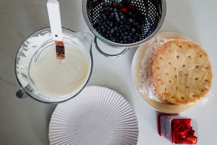 A mixing bowl of creamy tres leches batter, fresh blueberries in a colander, wrapped sponge cakes on a plate, some sliced strawberries in a dish, and an empty white plate—all laid out on a white surface.
