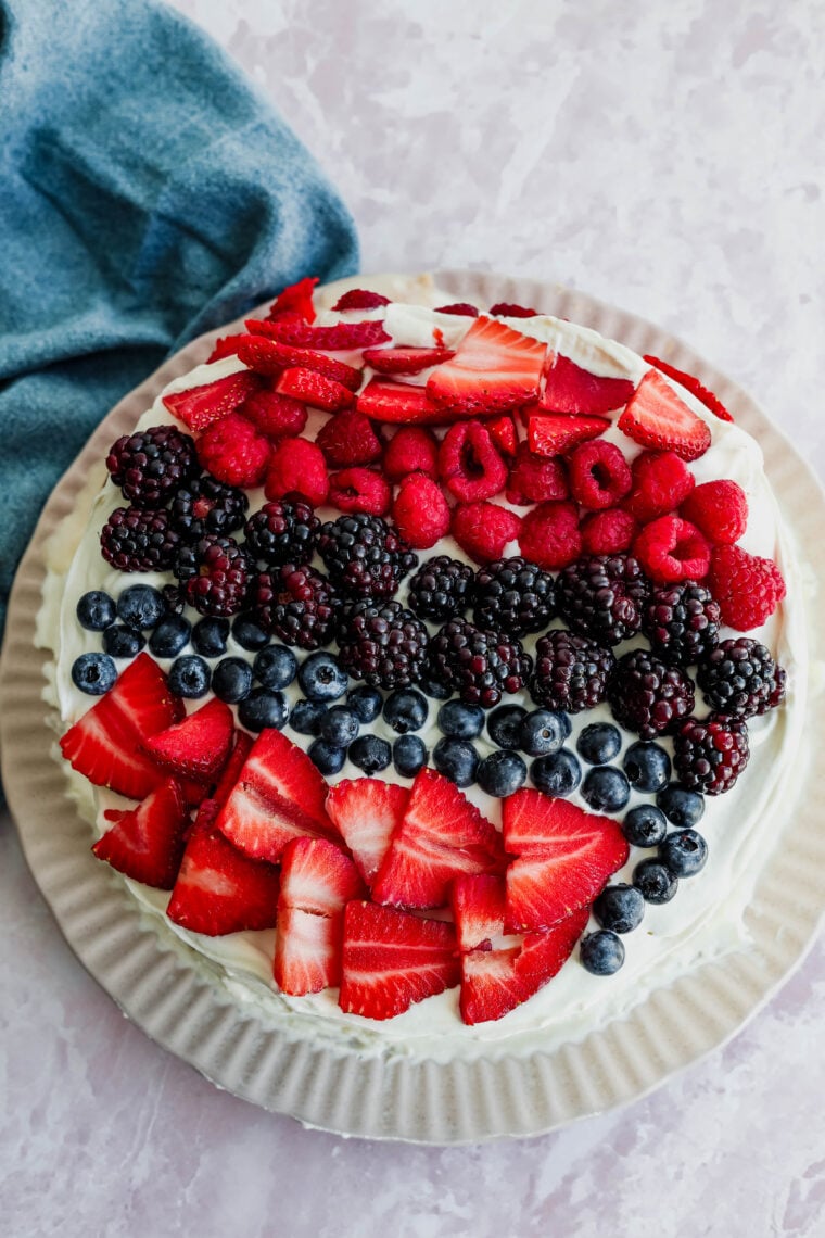 This round tres leches cake is covered in white frosting and loaded with rows of fresh strawberries, raspberries, blackberries, and blueberries. Its sitting on a light plate with a blue cloth close by.