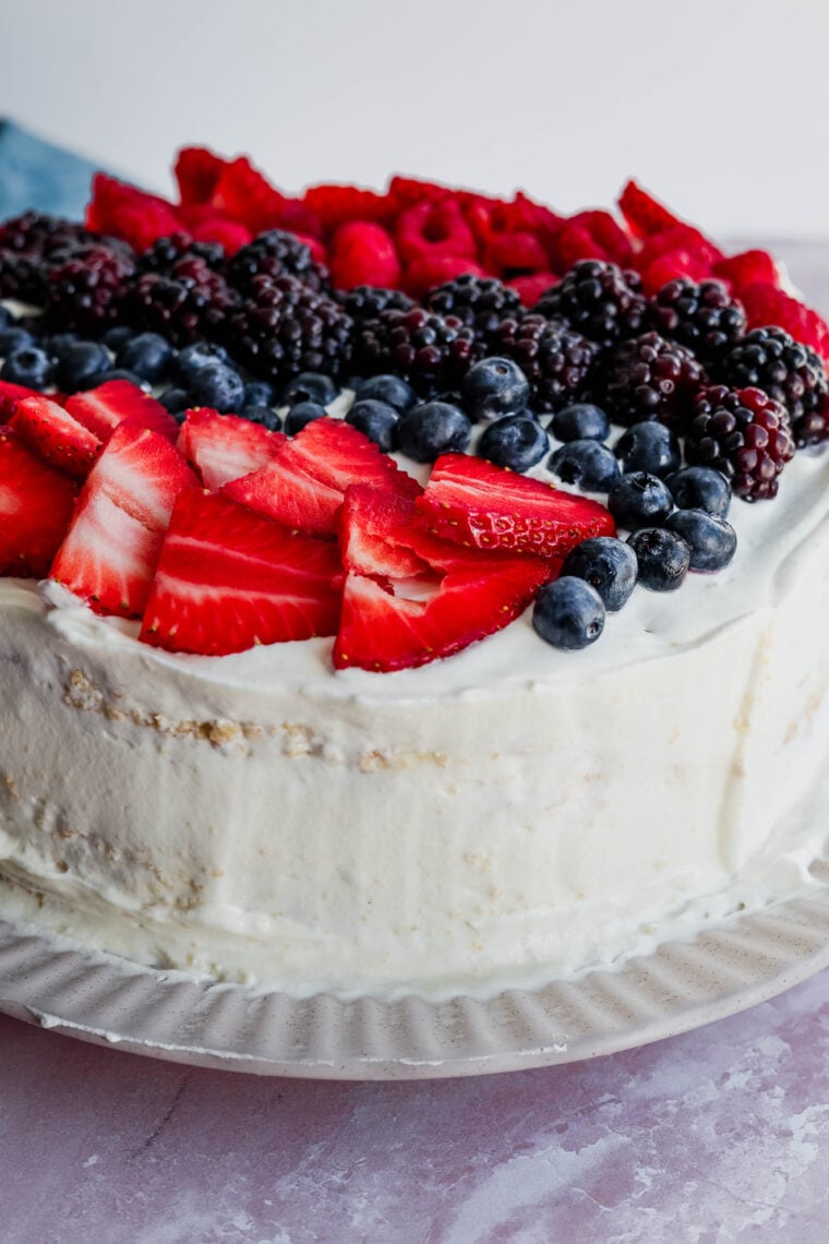 A round tres leches cake with white frosting, topped with fresh strawberries, blueberries, blackberries, and raspberries—it’s all set on a white plate and looks super delicious!.