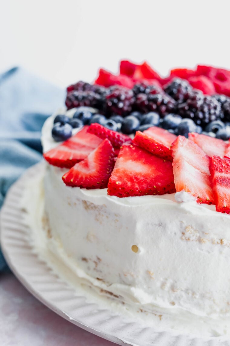 A close-up shot of a tres leches cake covered in white frosting and topped with sliced strawberries, whole blueberries, and blackberries. It’s sitting on a white plate with a blue cloth in the background.