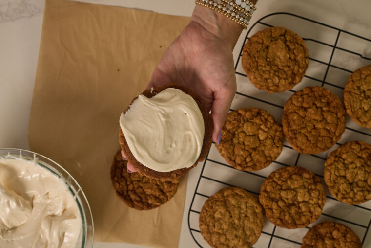 Someone’s holding up a browned butter oatmeal cream pies with a drizzle of white icing, right above a cooling rack stacked with plain cookies. There’s also a bowl of icing and some parchment paper nearby.