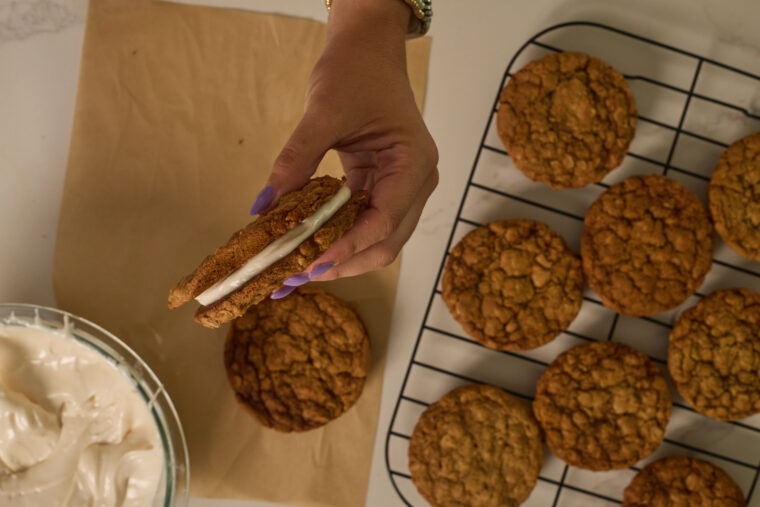 Someone’s holding up two browned butter oatmeal cream pies filled with white frosting. In the background, you can see more cookies chilling on a cooling rack and some parchment paper, plus there’s a bowl of extra frosting close by.
