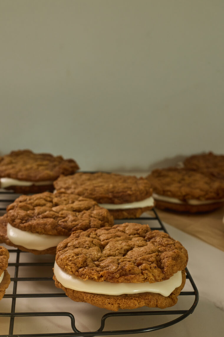 There are four browned butter oatmeal cream pies with white filling chilling on a black wire rack, and you can spot more in the background on a wooden table, all up against a plain beige wall.
