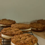 There are four oatmeal cookie pies with white filling chilling on a black wire rack, and you can spot more in the background on a wooden table, all up against a plain beige wall.