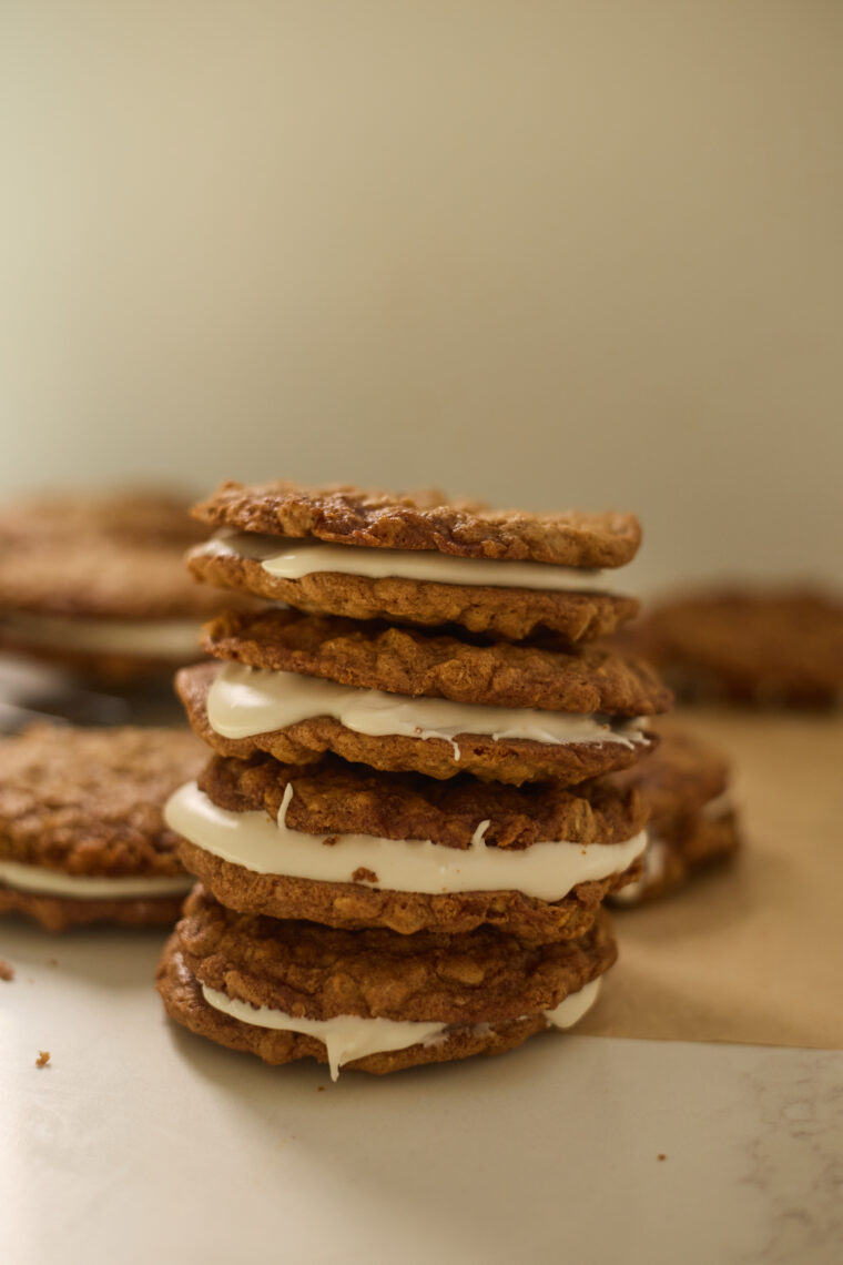 A stack of browned butter oatmeal cookie pies stuffed with white cream frosting sits on parchment paper, and you can see more cookies hanging out in the blurry background.