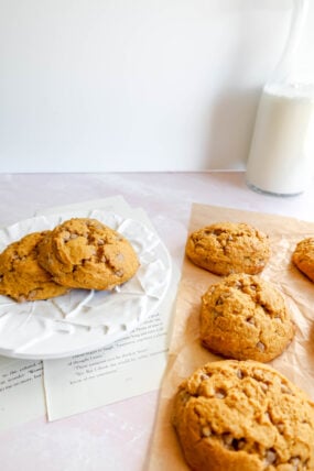 Pumpkin cinnamon cookies sit on a white plate and parchment paper next to a milk-filled glass bottle. Soft lighting adds coziness, with book pages peeking from under the plate.