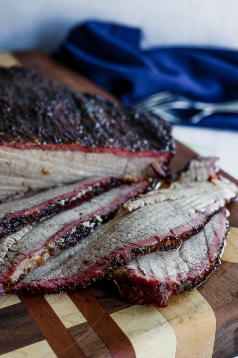 Sliced smoked brisket on a wooden board, showing off its juicy inside and tasty dark crust with a hint of smoke ring. There’s a blue cloth and some utensils chilling in the blurred background.