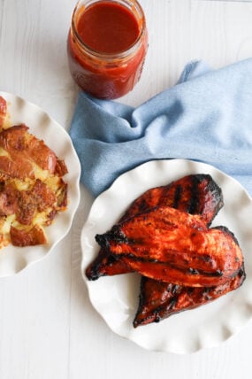 Smoked BBQ chicken breasts with roasted potatoes served up on a white plate—plus a jar of BBQ sauce and a blue cloth in the background for that extra vibe.