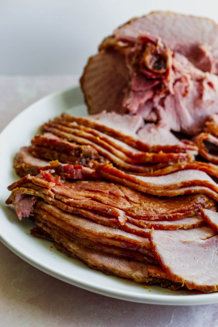 Sliced smoked glazed ham piled on a plate, with a big bone-in chunk in the back—super juicy, tender meat and that awesome caramelized crust.