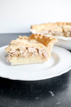 A slice of meat pie sits on a white plate, showing off its golden crust and tender filling. Its perfect for Thanksgiving dinner! In the background, theres more pie in the dish, waiting on a dark surface and tempting you to dig into this tasty homemade treat.