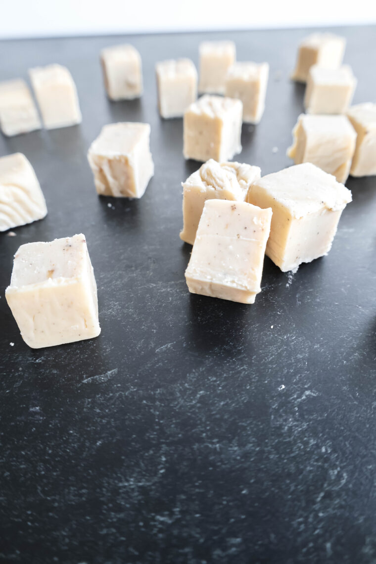 Chunks of white fudge made with browned butter and white chocolate are scattered around on a dark, textured surface. Theyre all slightly different sizes and have this creamy look with a shiny touch. Some pieces blur into the background for that artsy vibe.