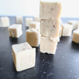 A bunch of creamy white fudge cubes made with yummy white chocolate are stacked up on a dark surface, with a few pieces teasing you from the background.