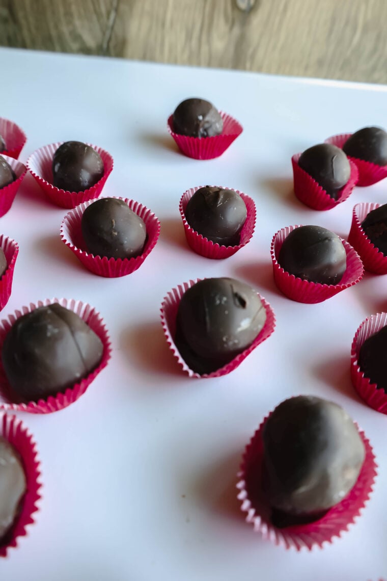 Chocolate truffles, just like cute candy treats, sit in red paper cups on a white backdrop. The whole scene has a wooden vibe in the background. These tasty bites come in different sizes and are scattered around randomly, making for an irresistibly tempting sight!.