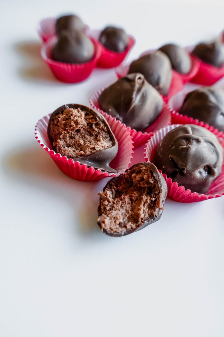 Chocolate truffles are chilling in red paper cups on a white background. Ones cracked open, showing off its rich buttercream middle. Theyre all wrapped up in silky dark chocolate and look super tempting!.