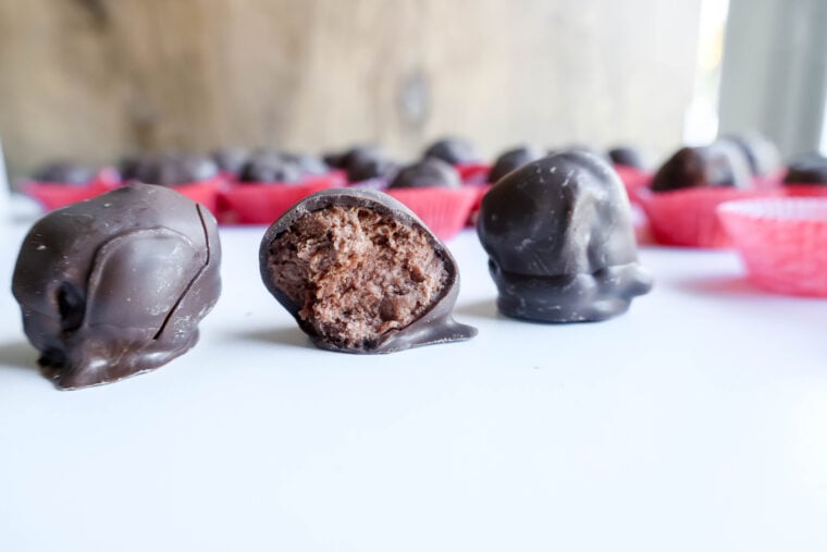 A bunch of chocolate truffles chilling on a white surface. Ones sliced open, showing off its yummy buttercream center. In the background, you can see blurry red paper cups teasing at more treats. Total indulgence vibes!.