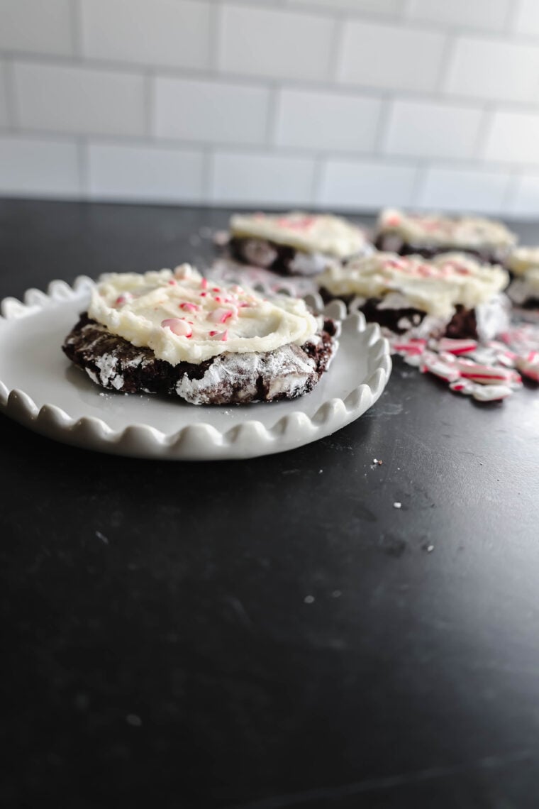 Theres a yummy chocolate peppermint cookie with white frosting and red sprinkles chilling on a fancy-edged white plate. You can spot more cookies kind of blurry in the back, sitting on a dark counter with a white tile wall looking classy behind them.
