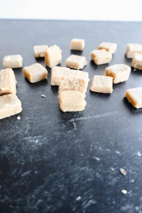Chunks of creamy maple fudge are spread out on a dark surface, with one piece stacked on another showing off a bite and its smooth, slightly grainy inside. This easy 3-ingredient recipe adds indulgent sweetness to any treat platter.