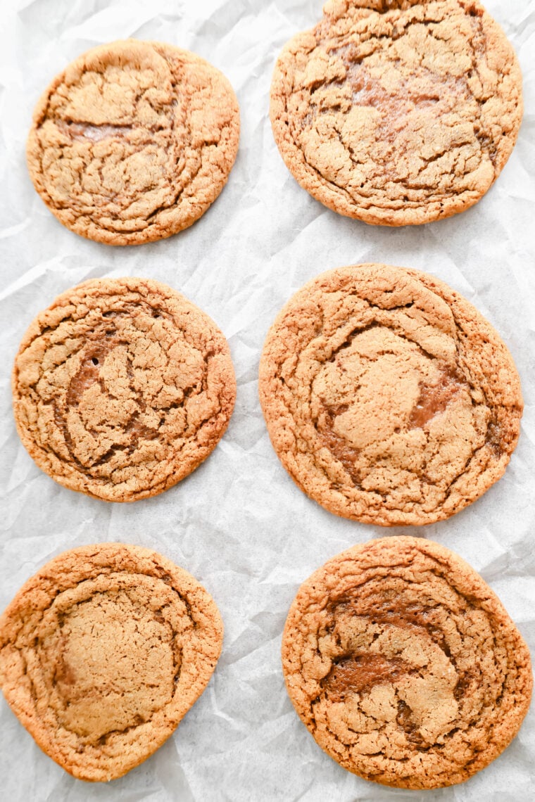 Six homemade molasses cookies are chilling on some crinkled parchment paper, looking soft and chewy. They’re golden brown with cool darker swirls, and have that slightly cracked surface that just screams perfect texture!.