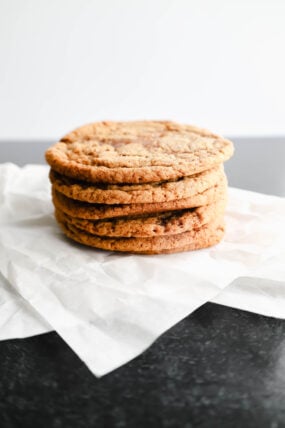 A pile of five yummy golden brown cookies, kind of like chewy molasses goodies, sits on white parchment paper. Little chocolate chunks pop out from the soft texture, all laid out nicely on a dark, slightly shiny surface.