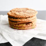 A pile of five yummy golden brown cookies, kind of like chewy molasses goodies, sits on white parchment paper. Little chocolate chunks pop out from the soft texture, all laid out nicely on a dark, slightly shiny surface.