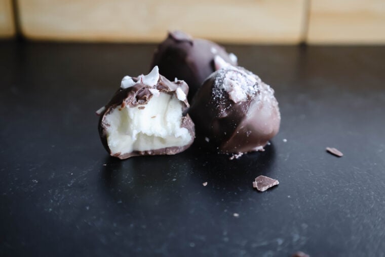 Three chocolate truffles chill out on a dark background—ones already had a nibble taken, showing off its creamy white buttercream insides. Little chocolate bits are sprinkled around, making the scene super tempting for anyone with a sweet tooth!.