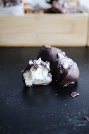 Three yummy chocolate truffles chill on a dark surface, with one showing off its creamy white filling. In the blurry background, theres a wooden box teasing more goodies. These treats totally remind you of festively indulgent peppermint buttercream candies—perfect for sprucing up your dessert game!.