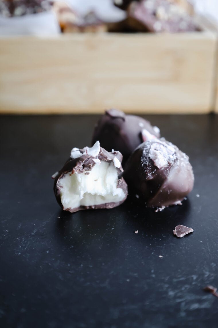Three yummy chocolate truffles chill on a dark surface, with one showing off its creamy white filling. In the blurry background, theres a wooden box teasing more goodies. These treats totally remind you of festively indulgent peppermint buttercream candies—perfect for sprucing up your dessert game!.