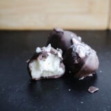 Three yummy chocolate truffles chill on a dark surface, with one showing off its creamy white filling. In the blurry background, theres a wooden box teasing more goodies. These treats totally remind you of festively indulgent peppermint buttercream candies—perfect for sprucing up your dessert game!.