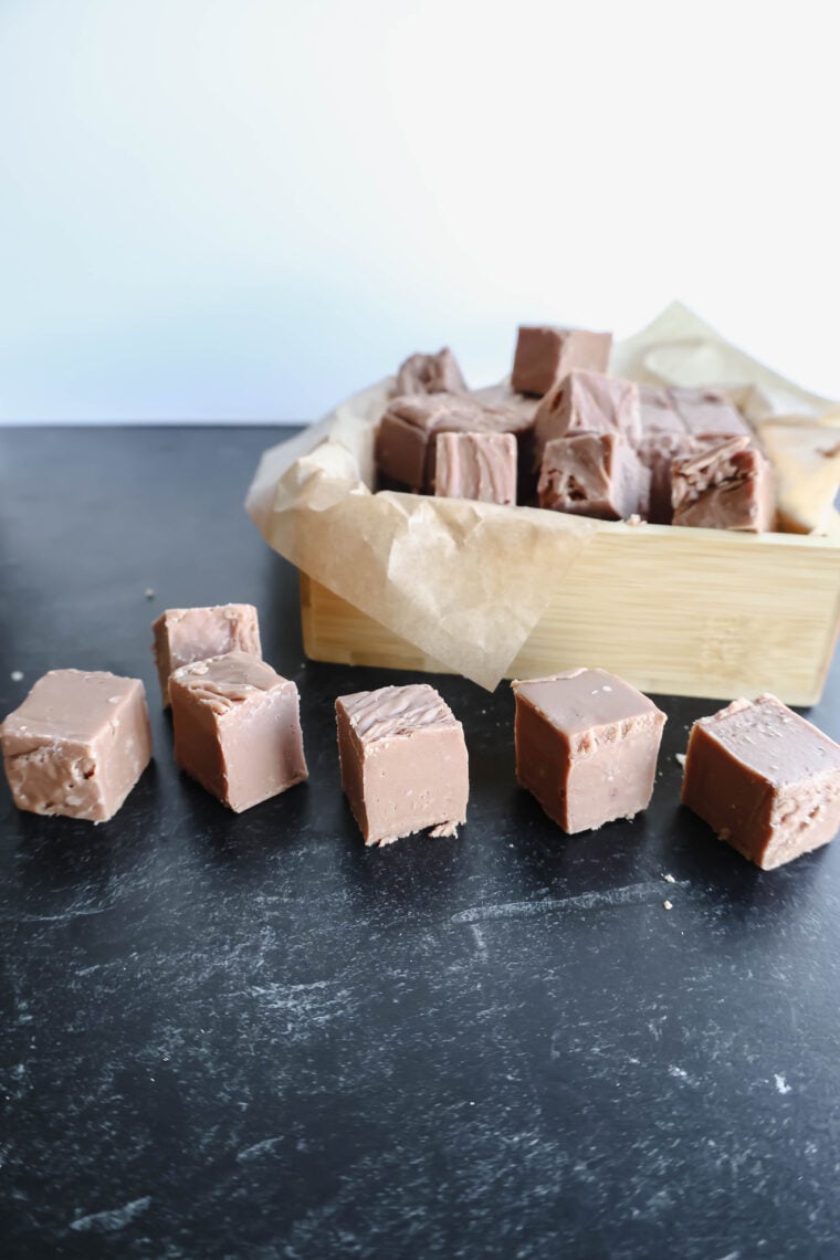 Chunks of milk chocolate fudge are scattered on a dark surface, and you can spot more pieces chilling in a wooden box with parchment paper. This Sees Copycat fudge has an ultra-smooth and rich vibe going on.