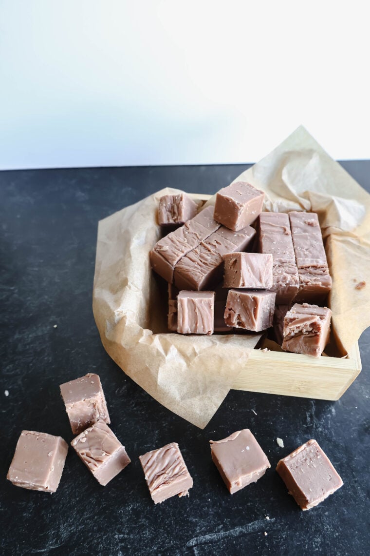 A wooden box lined with brown paper is filled with chunks of Copycat Milk Chocolate Fudge. A few pieces are scattered on the dark countertop, flaunting their smooth, rich texture.