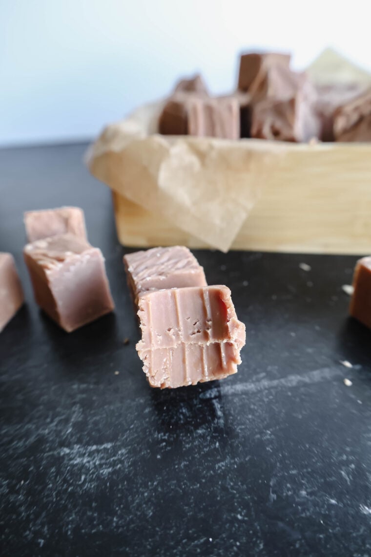 Heres a close-up of some silky milk chocolate fudge on a dark table. In the back, there’s a wooden tray with parchment paper loaded with more yummy Sees Fudge-style goodies, some with tempting little bite marks!.