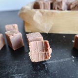 Heres a close-up of some silky milk chocolate fudge on a dark table. In the back, there’s a wooden tray with parchment paper loaded with more yummy Sees Fudge-style goodies, some with tempting little bite marks!.