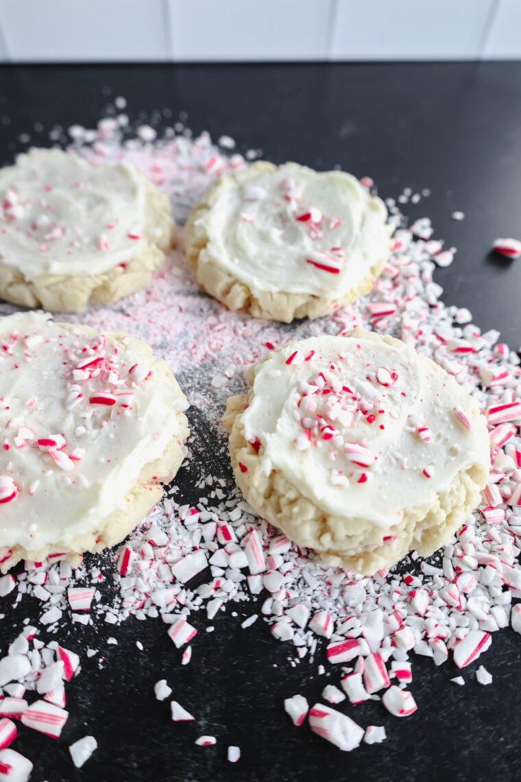 Four sugar cookies with white frosting are topped with crushed candy canes, creating a festive holiday treat. More crushed candy canes are scattered on the dark surface around the cookies. Alright, picture this: four sugar cookies with white frosting piled high with crushed candy canes. Perfect little bites of holiday cheer! Oh, and there are more crushed candy canes sprinkled all over, adding to the festive vibe.