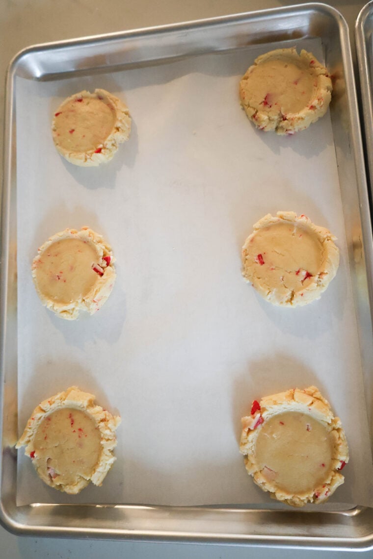 On a sheet lined with parchment paper, there are six unbaked sugar cookie dough balls, each dotted with red candy cane bits. Theyre spread out evenly and bring some festive holiday vibes to the baking scene.