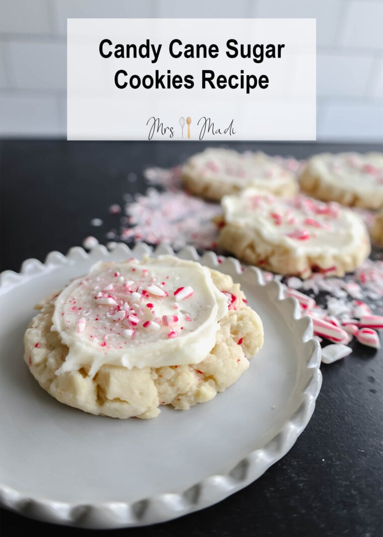 Youve got a holiday sugar cookie covered in white frosting with crushed candy canes on top, sitting on a plate. More cookies and candy cane bits are scattered around on a dark background. The words Candy Cane Sugar Cookies Recipe are cheerfully displayed above.