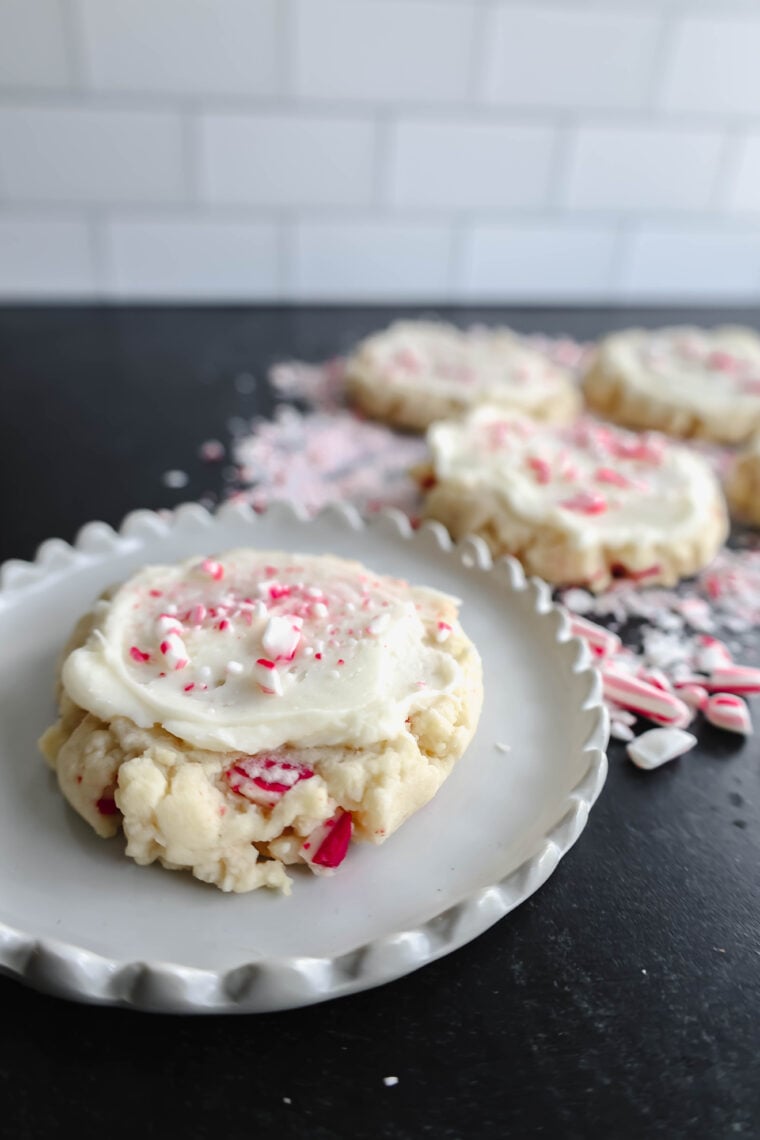 A frosted sugar cookie with crushed peppermint candy is front and center, bringing those holiday vibes. Behind it, theres a bunch of similar cookies and scattered candies giving off a festive candy cane wonderland feel on a black surface.