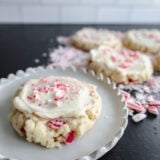 A frosted sugar cookie with crushed peppermint candy is front and center, bringing those holiday vibes. Behind it, theres a bunch of similar cookies and scattered candies giving off a festive candy cane wonderland feel on a black surface.
