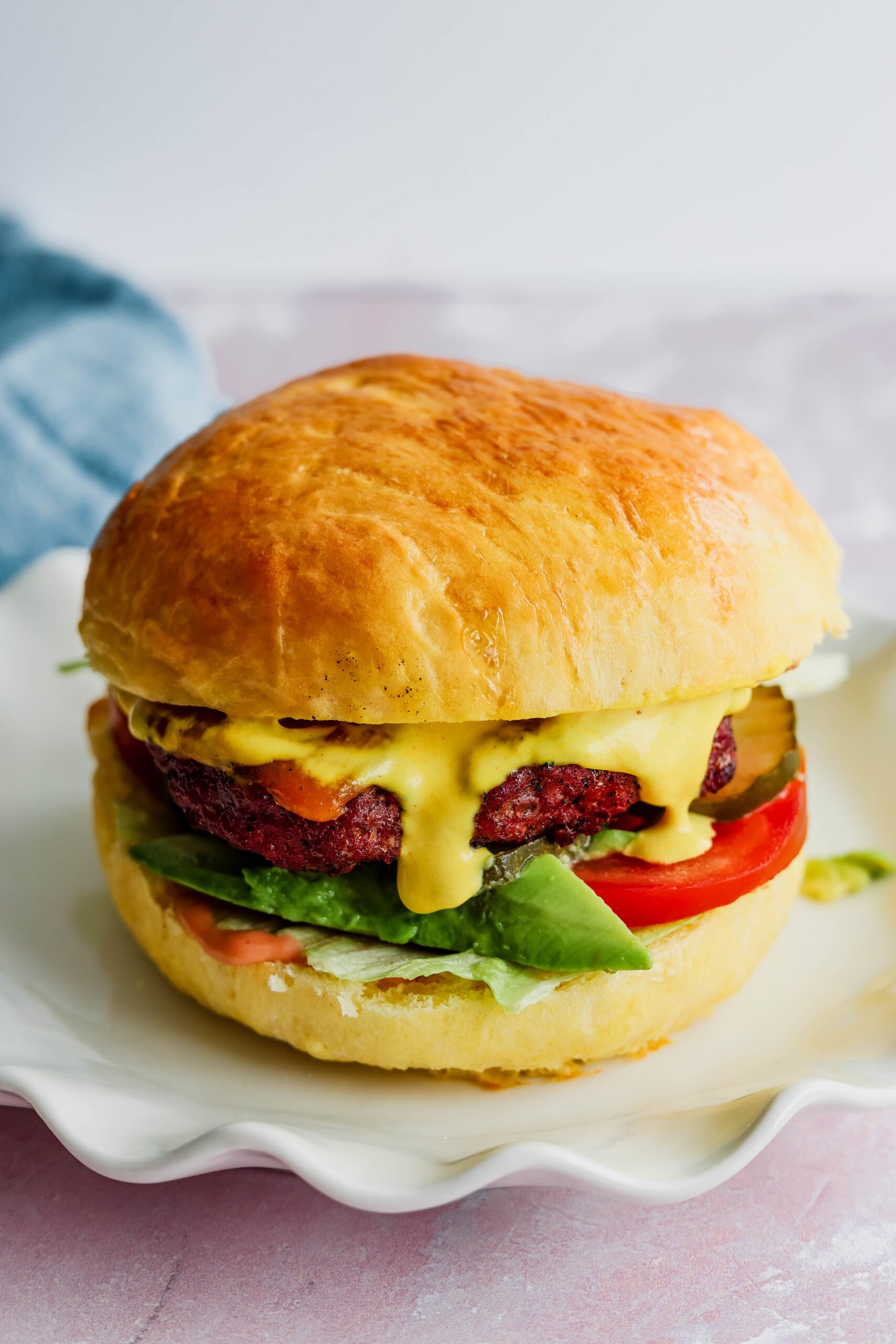Check out this juicy American burger—golden bun, fresh greens, tomato, pickles, a tasty patty, and gooey melted cheese sauce. It’s all stacked up on a white plate with a blue napkin in the background.