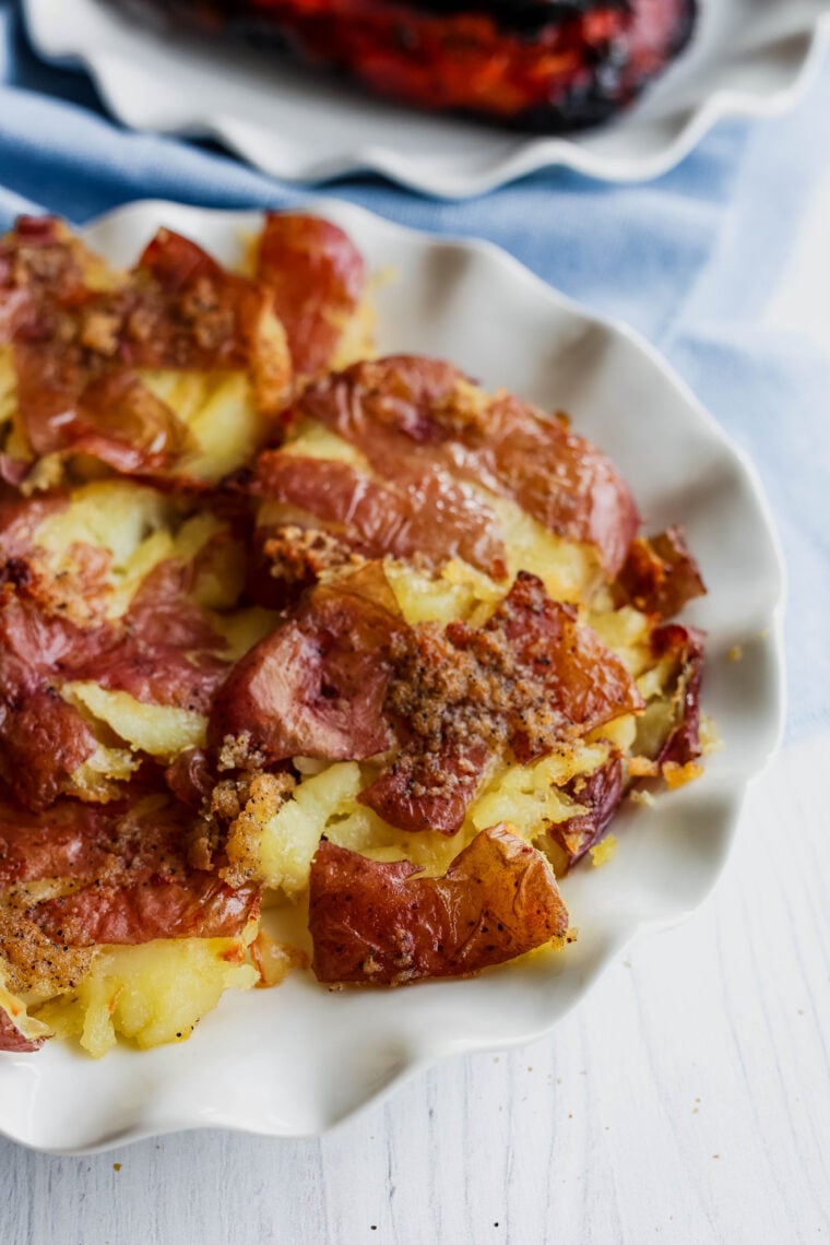 A plate of crispy smashed potatoes, baked to perfection and loaded with seasoning, is sitting on a white table next to a blue napkin. You can see another dish peeking out in the background.