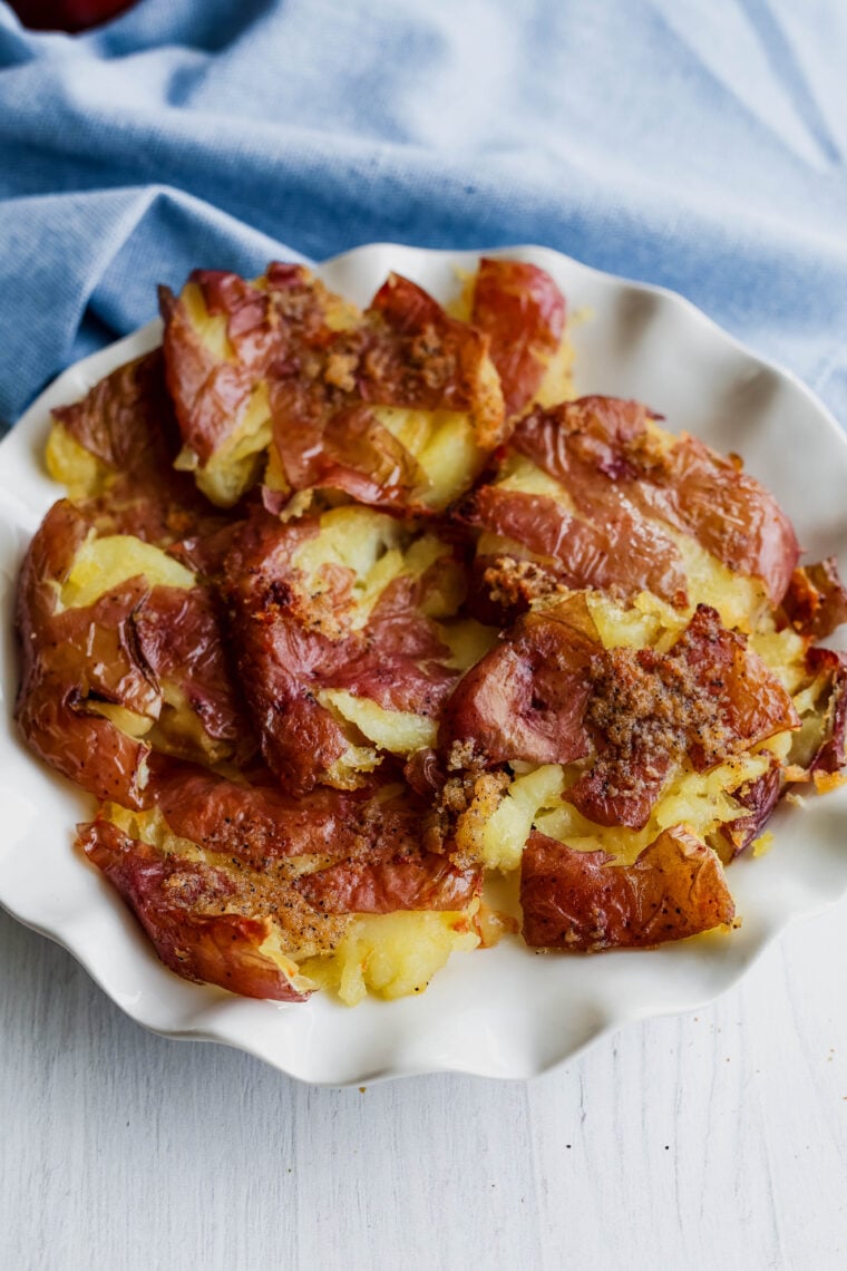 A white plate loaded with crispy smashed potatoes, sprinkled with golden seasoning, is set on a white surface. Theres a light blue cloth hanging out in the background.